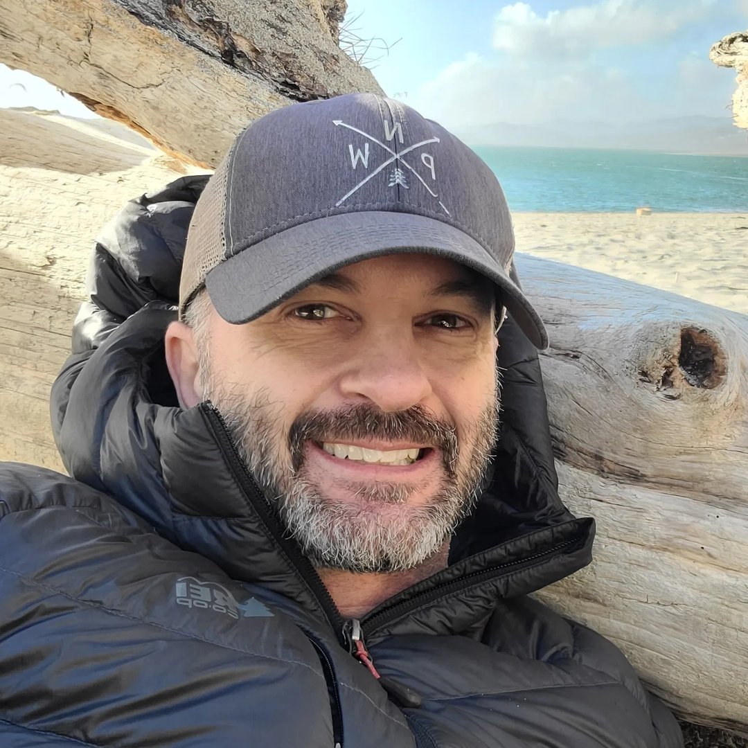 Jeremy Henricks in a PNW cap, leaning on driftwood on an Oregon beach